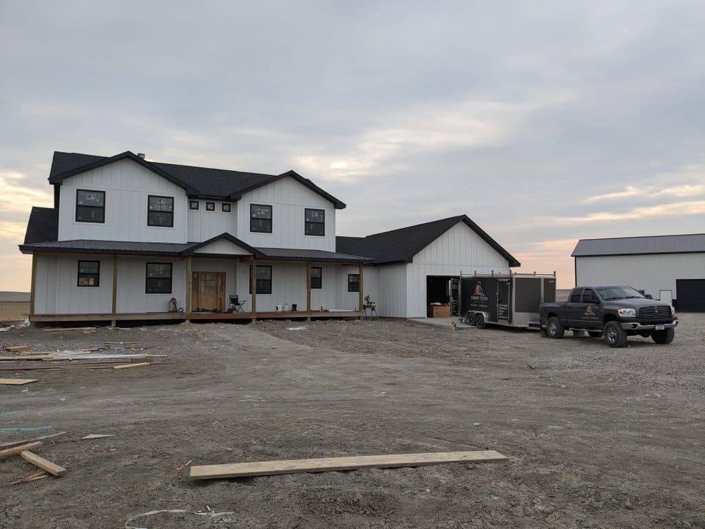 New Construction Farmhouse Style Home With Truck Newly built farmhouse with black roof, white siding, truck, and trailer on a gravel lot.